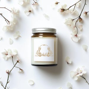 May include: A brown glass candle jar with a silver lid and a white label with a gold floral design. The candle is surrounded by white flower petals.