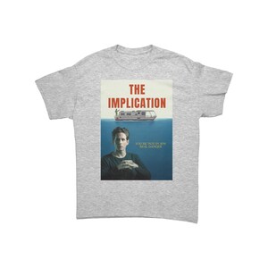 May include: A light grey t-shirt with a graphic from the show "It's Always Sunny in Philadelphia." The graphic features the words "THE IMPLICATION" above a boat on water, and a person below the water line. The text "YOU'RE NOT IN ANY REAL DANGER" is also included.