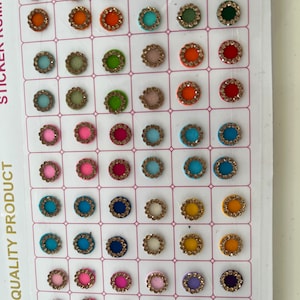 May include: A sheet of colourful round stickers with a gold rim and rhinestones. The stickers are arranged in a grid pattern. The text "A QUALITY PRODUCT STICKER KUMKUM" is printed on the sheet.