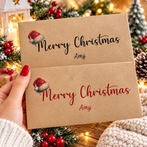 May include: Two rectangular kraft paper envelopes featuring a printed red and white Santa hat icon. The top envelope displays the text Merry Christmas and the name Amy in black ink. The bottom envelope features the same text in red ink.