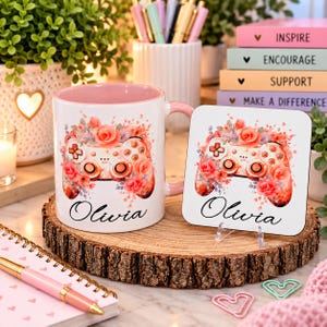 May include: A white ceramic mug with a pink interior and a matching square coaster, both featuring a floral game controller graphic and the name Olivia in black script. The mug has a pink handle and the coaster is displayed on a small clear stand.