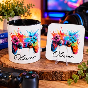 May include: A white ceramic mug and a matching square coaster, both featuring a colourful paint splatter design of a video game controller and the name Oliver in black script. The mug has a black interior and handle, while the coaster is white.
