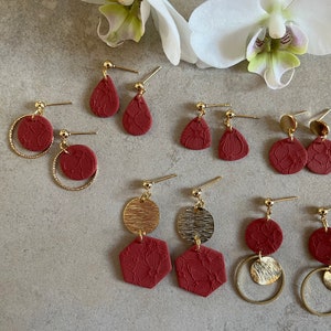 Könnte beinhalten: Eine Sammlung handgefertigter Ohrringe in verschiedenen Formen und Designs. Die Ohrringe haben einen tiefroten Farbton mit floralen Mustern und goldfarbenen Akzenten. Zu den Stilen gehören Tropfen-, Kreis- und geometrische Formen mit goldfarbenen Ohrhaken.