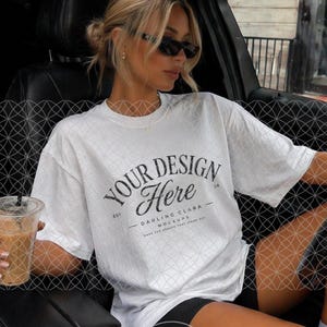 May include: White oversized t-shirt with the text "YOUR DESIGN Here" in black script. The shirt is paired with black shorts. The image is taken inside a car.