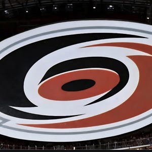 May include: Large, oval-shaped logo of the Carolina Hurricanes hockey team. The logo features a red, black, and white colour scheme with a stylized hurricane symbol at the centre. The logo is displayed in an arena setting.