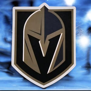 May include: A shield-shaped emblem featuring the logo of the Vegas Golden Knights hockey team. The logo is a stylized knight's helmet in gold and black, with a white border. The background is a blurred blue.