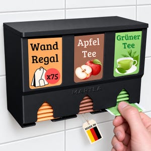 Op de afbeelding: Een zwarte wand-theezakjesdispenser met drie compartimenten met de labels Wand Regal, Apfel Tee en Gr&uuml;ner Tee. De unit is voorzien van het Martea-logo, een afbeelding van een theezakje met x75, een appelillustratie en een icoon van een kopje groene thee.
