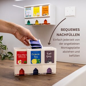 Op de afbeelding: Een witte theezakjeshouder van kunststof met drie verticale vakken voor het organiseren van theezakjes. De unit bevat labels voor Rose Hip & Hibiscus, Lemon Tea en Earl Grey. Inclusief een afneembare montageplaat voor wandmontage.