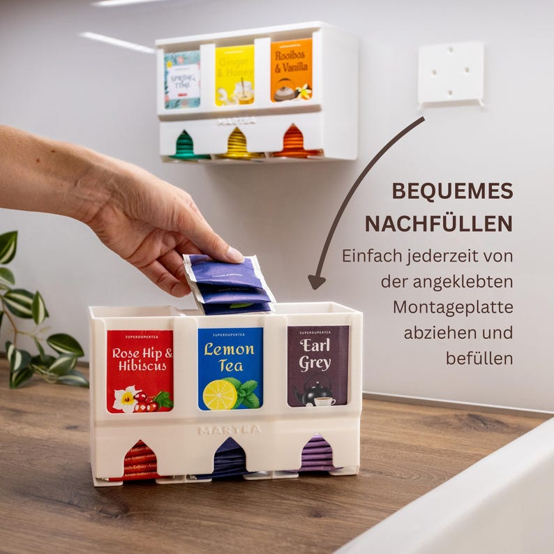 Op de afbeelding: Een witte theezakjeshouder van kunststof met drie verticale vakken voor het organiseren van theezakjes. De unit bevat labels voor Rose Hip & Hibiscus, Lemon Tea en Earl Grey. Inclusief een afneembare montageplaat voor wandmontage.