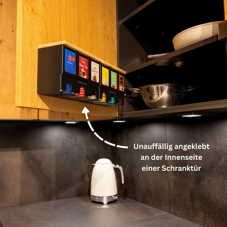 Op de afbeelding: Een zwarte theezakjesdispenser bevestigd aan de binnenkant van een houten kastdeur. De unit heeft vijf verticale sleuven, elk met een kleurrijke theedoos met zichtbare labels. Een witte waterkoker staat op het donkere aanrecht onder de kast.