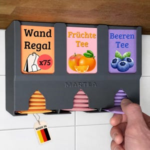 Op de afbeelding: Een donkergrijze wand-theezakjeshouder met het opschrift MARTEA en drie vakken. De labels bevatten de teksten Wand Regal, Fr&uuml;chte Tee en Beeren Tee, met illustraties van een theezakje, sinaasappel en bosbessen. Een label met de Duitse vlag is bevestigd.