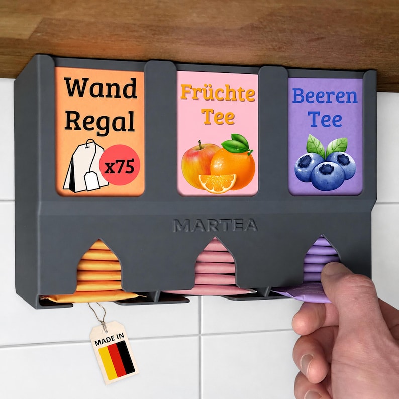 Op de afbeelding: Een donkergrijze wand-theezakjeshouder met het opschrift MARTEA en drie vakken. De labels bevatten de teksten Wand Regal, Fr&uuml;chte Tee en Beeren Tee, met illustraties van een theezakje, sinaasappel en bosbessen. Een label met de Duitse vlag is bevestigd.
