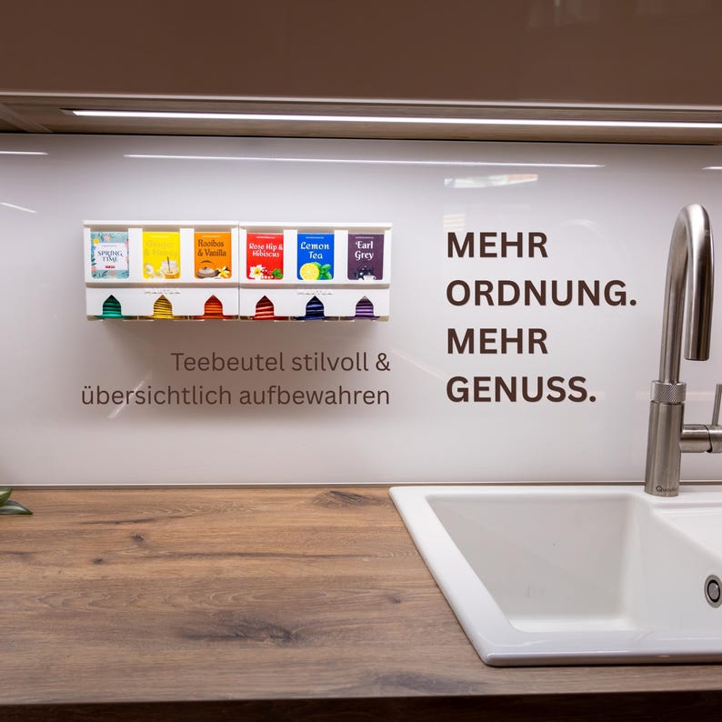 Op de afbeelding: Een witte wand-theezakjeshouder met zes kleurrijke theedoosjes. De unit bevat de Duitse tekst: MEHR ORDNUNG. MEHR GENUSS. Teebeutel stilvoll & &uuml;bersichtlich aufbewahren. Het is geplaatst naast een roestvrijstalen keukenkraan.