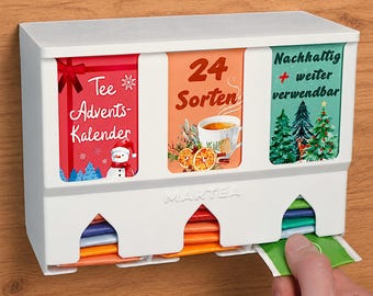 Op de afbeelding: Een witte plastic thee-adventskalenderdispenser met drie verticale vakken. De panelen zijn voorzien van feestelijke afbeeldingen, een sneeuwpop, een theekop en kerstbomen met Duitse tekst. De dispenser bevat theezakjes.