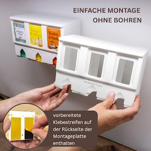 Op de afbeelding: Een witte theezakjeshouder van kunststof met drie verticale sleuven en de merknaam MARTEA in reli&euml;f op de voorzijde. De tekst EINFACHE MONTAGE OHNE BOHREN is bovenaan zichtbaar. De achterkant bevat gele plakstrips voor wandmontage.