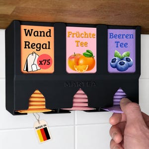 Op de afbeelding: Een zwarte wand-theezakjeshouder met drie verticale vakken. De labels tonen Wand Regal x75, Fr&uuml;chte Tee met een appel en sinaasappel, en Beeren Tee met bosbessen. De merknaam MARTEA is in reli&euml;f op de voorzijde aangebracht.