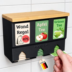 Könnte beinhalten: Ein schwarzer Wand-Teebeutelspender mit einer hellen Holzoberseite. Er verfügt über drei Fächer mit den Beschriftungen Wand Regal, Apfel Tee und Grüner Tee. Das Gerät ist mit MARTEA gekennzeichnet und enthält ein kleines Etikett in den Farben der deutschen Flagge.