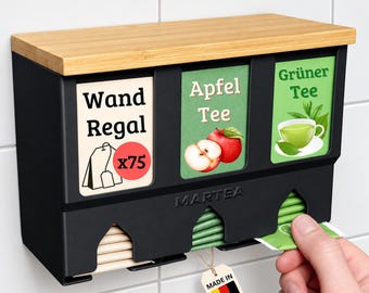 Może przedstawiać: Czarny ścienny dozownik do torebek herbaty z jasnym drewnianym blatem. Posiada trzy przegródki z etykietami Wand Regal, Apfel Tee oraz Grüner Tee. Urządzenie jest oznaczone marką MARTEA i zawiera małą zawieszkę w barwach flagi niemieckiej.