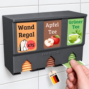Op de afbeelding: Een donkergrijze theezakjeshouder voor aan de muur met drie vakken. De labels bevatten de teksten Wand Regal, Apfel Tee en Gr&uuml;ner Tee. De houder toont afbeeldingen van een theezakje, een appel en een kop groene thee. De merknaam MARTEA staat op de voorzijde.