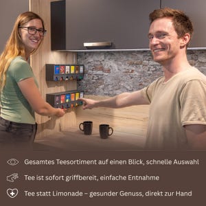 Op de afbeelding: Twee aan de muur gemonteerde donkergrijze theezakjesdispensers met meerdere vakken, elk met kleurrijke theedoosjes. De dispensers hebben een uitgifte-mechanisme aan de onderkant. Onderaan staat de tekst: Gesamtes Teesortiment auf einen Blick, schnelle Auswahl, Tee ist sofort griffbereit, einfache Entnahme, Tee statt Limonade &ndash; gesunder Genuss, direkt zur Hand.