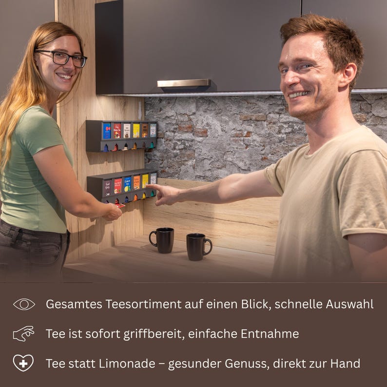 Op de afbeelding: Twee aan de muur gemonteerde donkergrijze theezakjesdispensers met meerdere vakken, elk met kleurrijke theedoosjes. De dispensers hebben een uitgifte-mechanisme aan de onderkant. Onderaan staat de tekst: Gesamtes Teesortiment auf einen Blick, schnelle Auswahl, Tee ist sofort griffbereit, einfache Entnahme, Tee statt Limonade &ndash; gesunder Genuss, direkt zur Hand.