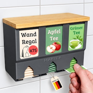 Op de afbeelding: Een donkergrijze wandtheezakjeshouder met een lichthouten bovenkant. Het heeft drie compartimenten met labels waarop Wand Regal, Apfel Tee en Gr&uuml;ner Tee staat. De unit is voorzien van de merknaam MARTEA en een label met de Duitse vlag.