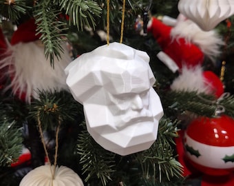 Op de afbeelding: Een witte, geometrische kerstversiering in de vorm van een buste van een persoon met een baard en snor. De versiering hangt aan een gouden draad aan een kerstboom met andere versieringen en groen.