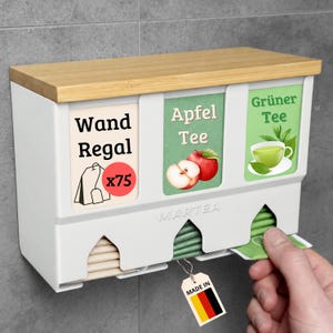 Op de afbeelding: Een witte theezakjeshouder voor aan de muur met een lichthouten bovenkant. De unit heeft drie vakken met de labels Wand Regal, Apfel Tee en Gr&uuml;ner Tee. De houder bevat theezakjes, waarvan er &eacute;&eacute;n uit de rechter opening wordt getrokken.