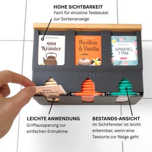 Op de afbeelding: Een donkergrijze theezakjeshouder met een bamboe bovenkant, voorzien van drie verticale vakken voor het tonen en doseren van theezakjes. De unit is gelabeld met MARTEA en bevat theezakjes met de labels 999 Kr&auml;uter, Rooibos & Vanilla en Spring Time.