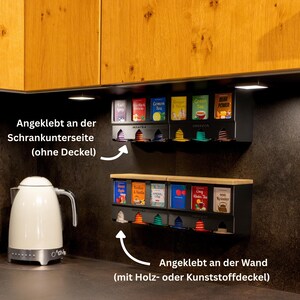 Op de afbeelding: Twee zwarte theezakjesdispensers gemonteerd in een keuken. De bovenste unit is onder een kast bevestigd en bevat zes theedoosjes. De onderste unit is aan de muur bevestigd met een houten bovenkant en bevat zes theedoosjes.