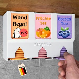 Op de afbeelding: Een witte wand-theezakjeshouder met drie compartimenten gelabeld Wand Regal, Fr&uuml;chte Tee en Beeren Tee. De unit is voorzien van het Martea-logo, afbeeldingen van fruit en theezakjes, en een klein label met de kleuren van de Duitse vlag.