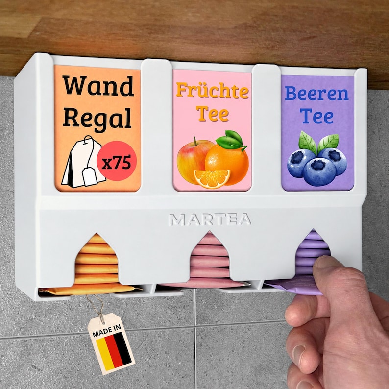 Op de afbeelding: Een witte wand-theezakjeshouder met drie compartimenten gelabeld Wand Regal, Fr&uuml;chte Tee en Beeren Tee. De unit is voorzien van het Martea-logo, afbeeldingen van fruit en theezakjes, en een klein label met de kleuren van de Duitse vlag.