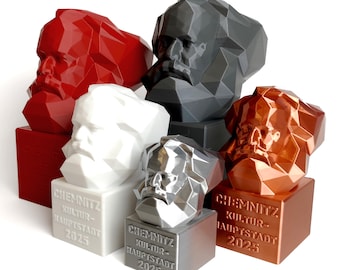 Op de afbeelding: Vijf 3D-geprinte bustes van Karl Marx in verschillende kleuren: rood, wit, zilver, grijs en koper. Elk buste staat op een vierkante basis met de tekst "Chemnitz Kultur-Hauptstadt 2025".