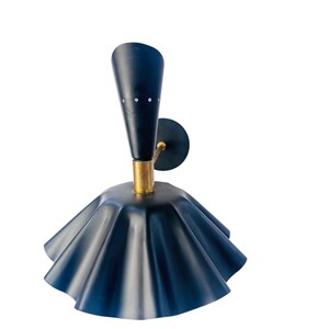 Op de afbeelding: Een marineblauwe wandlamp met een uniek ontwerp. De lamp heeft een uitlopende, gerimpelde kap en een conische bovenkant met drie kleine witte stippen. De basis is goudkleurig en de armatuur is gemonteerd op een cirkelvormige zwarte plaat.