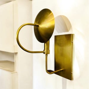 Könnte beinhalten: Eine Wandleuchte aus Messing mit rundem Schirm und rechteckiger Grundplatte. Die Leuchte hat einen gebogenen Arm, der sich von der Grundplatte erstreckt, um den Schirm zu tragen. Die Oberfläche ist ein warmes, metallisches Gold. Das Design ist modern und minimalistisch.