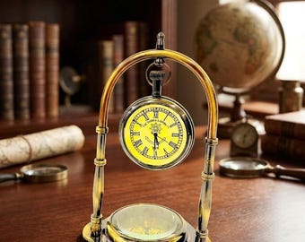 May include: A brass-toned metal desk clock suspended within an arched frame, featuring a yellow clock face with black Roman numerals and the text "VICTORIA LONDON". A circular compass is integrated into the base of the stand.
