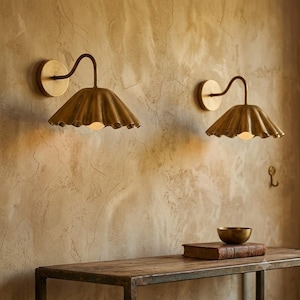Peut inclure: Deux appliques murales en laiton avec des bras incurvés et des abat-jours larges, festonnés et en forme de cloche. Chaque luminaire possède une fixation murale circulaire et une source lumineuse orientée vers le bas, diffusant une lueur chaude.