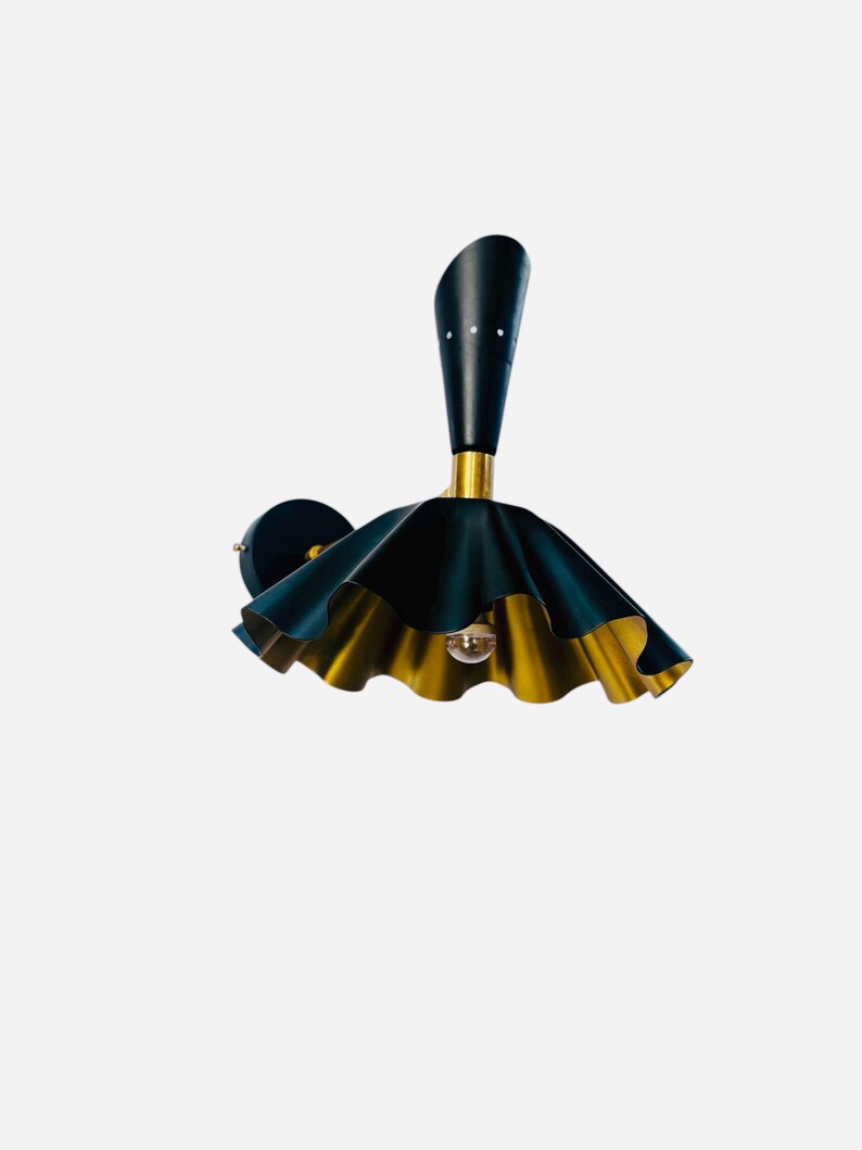Op de afbeelding: Een wandlamp met een zwart en goud kleurenschema. De lamp heeft een langwerpige zwarte bovenkant met drie witte stippen, een gouden basis en een gefranjerde, parapluvormige kap met een zwarte buitenkant en een gouden binnenkant. Een decoratief verlichtingselement.