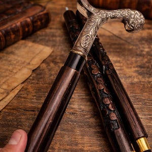 Könnte beinhalten: Nahaufnahme von drei antiken Spazierstöcken. Die Stöcke haben dunkelbraune Holzschäfte und verzierte silberfarbene Griffe mit floralen und scrollartigen Designs. Eine Gehhilfe wird von einer Hand gehalten. Der Hintergrund ist eine Holzoberfläche.