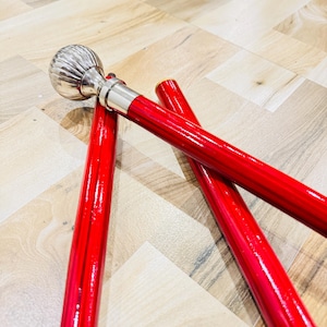 May include: A red walking cane with a silver-colored, ribbed ball-shaped handle. The cane's shaft is a vibrant red color. The cane is lying on a light-colored wooden floor.