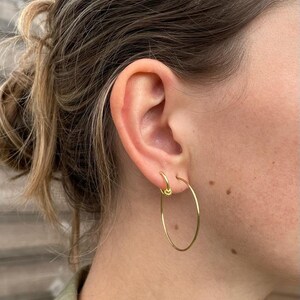 Op de afbeelding: Close-up van een oor met twee gouden oorringen. Eén is een kleine knop, de andere is een grote, dunne ring. De oorbellen zijn goudkleurig. De huidskleur is licht.