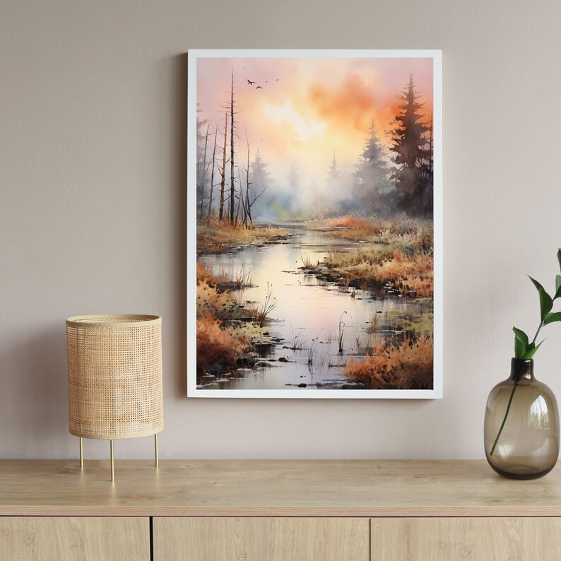 May include: A watercolor painting of a misty forest scene with a river winding through the trees. The sky is a soft orange and pink, and the trees are a muted green and brown. The painting is framed in a white frame.