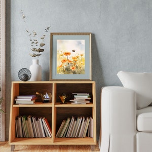 May include: A framed watercolor painting of orange daisies with a bee, set in a gold frame. The painting is displayed on a wooden bookshelf with books, a white vase, and decorative items. The background is a light blue wall.