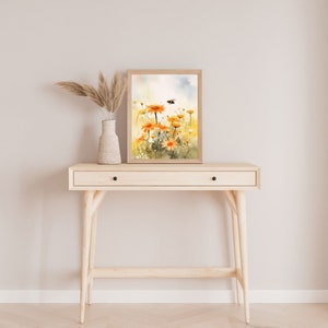 May include: A framed watercolor print of orange daisies with a bee, displayed on a light wood console table. A white vase with dried pampas grass sits to the left. The table has a single drawer and black knobs.