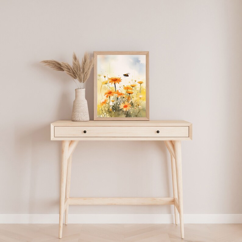 May include: A framed watercolor print of orange daisies with a bee, displayed on a light wood console table. A white vase with dried pampas grass sits to the left. The table has a single drawer and black knobs.