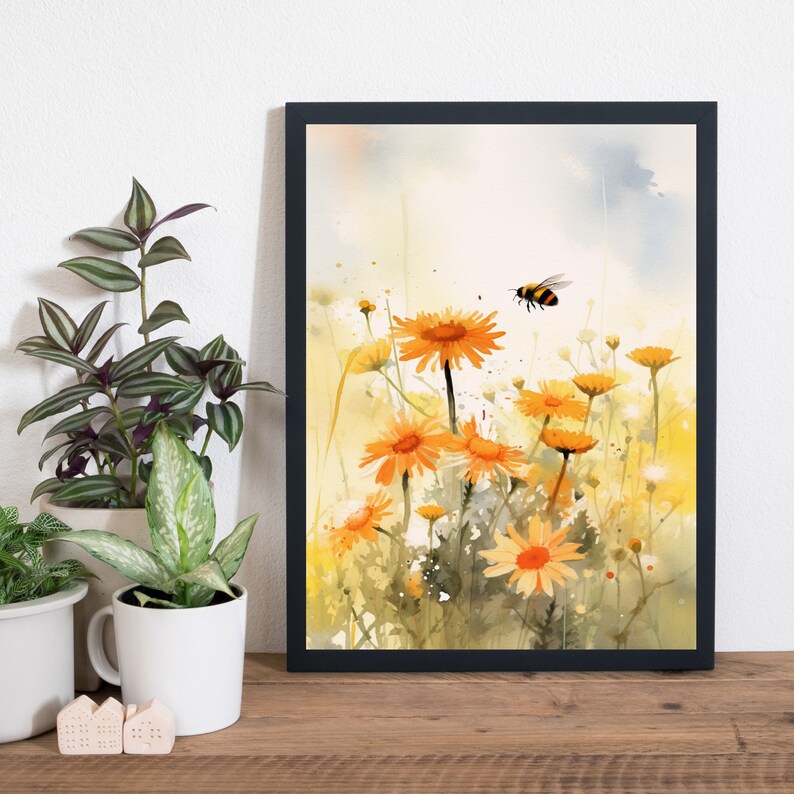 May include: A framed watercolor print featuring a field of orange daisies with a flying bee. The artwork has a soft, blurred style with yellow and green hues, set against a light blue sky. The frame is black, and the print is displayed on a wooden surface.