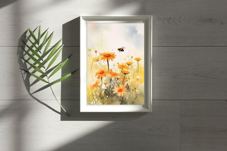 May include: A framed watercolor print featuring a field of orange daisies with a flying bee. The artwork has a soft, painterly style with yellow and green hues. The white frame sits on a light wood surface, with a green leaf in the corner.