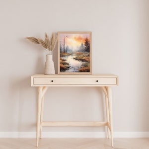 May include: A framed watercolor painting of a forest scene with a river running through it. The painting is in shades of brown, orange, and green, and it is set against a light beige wall. The painting is on top of a light wood console table with a drawer.