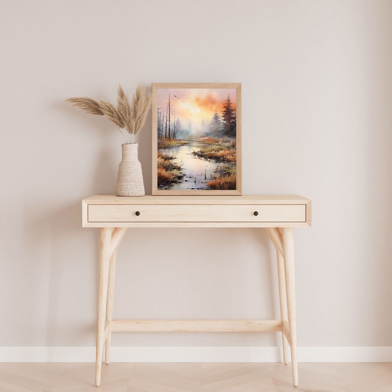 May include: A framed watercolor painting of a forest scene with a river running through it. The painting is in shades of brown, orange, and green, and it is set against a light beige wall. The painting is on top of a light wood console table with a drawer.