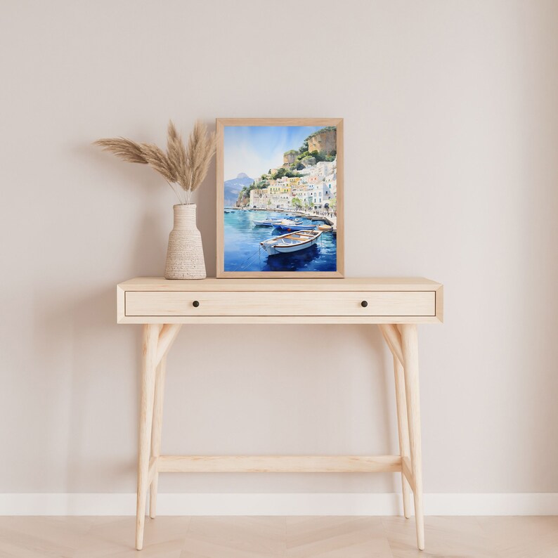 May include: A watercolor painting of a coastal town with colorful buildings and boats in a blue bay. The painting is framed in a light wood frame and is displayed on a light wood console table.